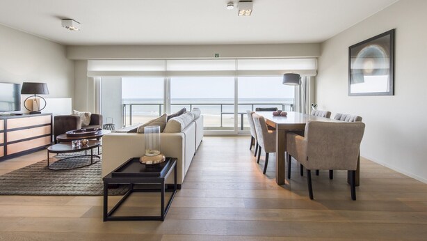 Ein geräumiges Wohnzimmer mit großem Fenster zum Strand, einem Essbereich mit Tisch und Stühlen sowie einer Sitzecke mit Sofa und Sesseln.