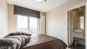 Ein Schlafzimmer mit einem großen Bett, einem Fenster mit Jalousien, einem Heizkörper und einem durch eine offene Tür erreichbaren Badezimmer.
