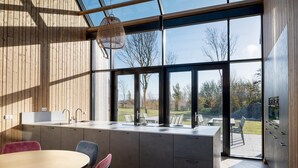 A modern kitchen with a large island, a dining area with chairs, and a view of the outdoors through large windows. - Holiday Home in Nieuwvliet-Bad near Beach (Nieuwvliet)