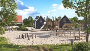 A playground with slides and climbing structures, surrounded by sand and greenery, with houses in the background. - Holiday Home in Nieuwvliet-Bad near Beach (Nieuwvliet)
