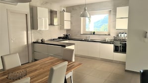 A modern kitchen with white cabinets, a wooden dining table, and a view of mountains through a window.
