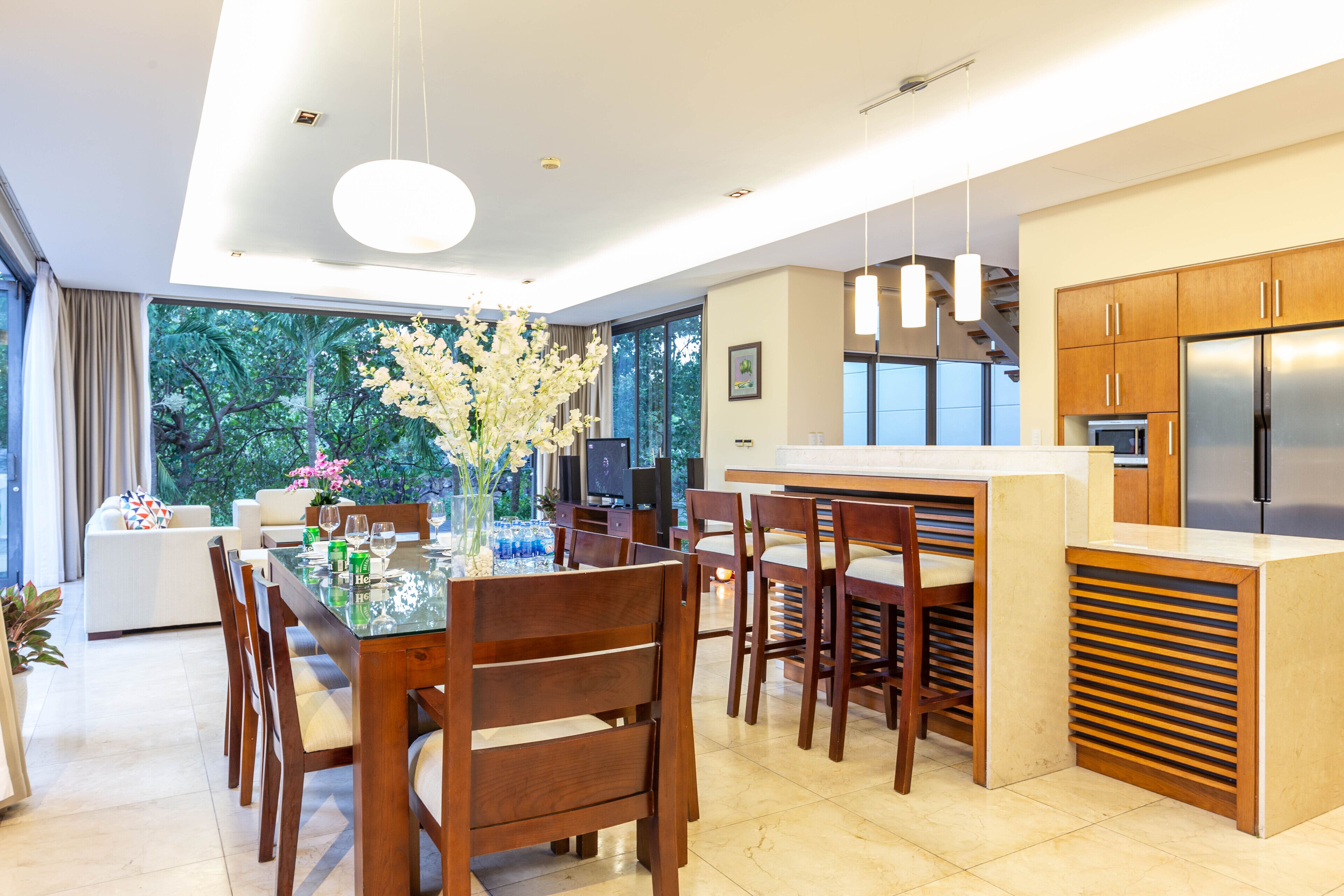 A modern kitchen with wooden cabinets, a stainless steel refrigerator, and a dining area with a glass table and wooden chairs.