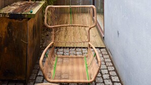 Ein Korbstuhl mit grünem Gestell steht auf einem Kopfsteinpflaster-Sitzplatz neben einem Holztisch und einer Mauer.