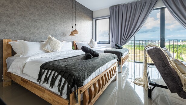 Une chambre moderne avec un grand lit, un lit plus petit et une chaise. Il y a une grande fenêtre avec des rideaux, un luminaire suspendu et une vue sur la ville.
