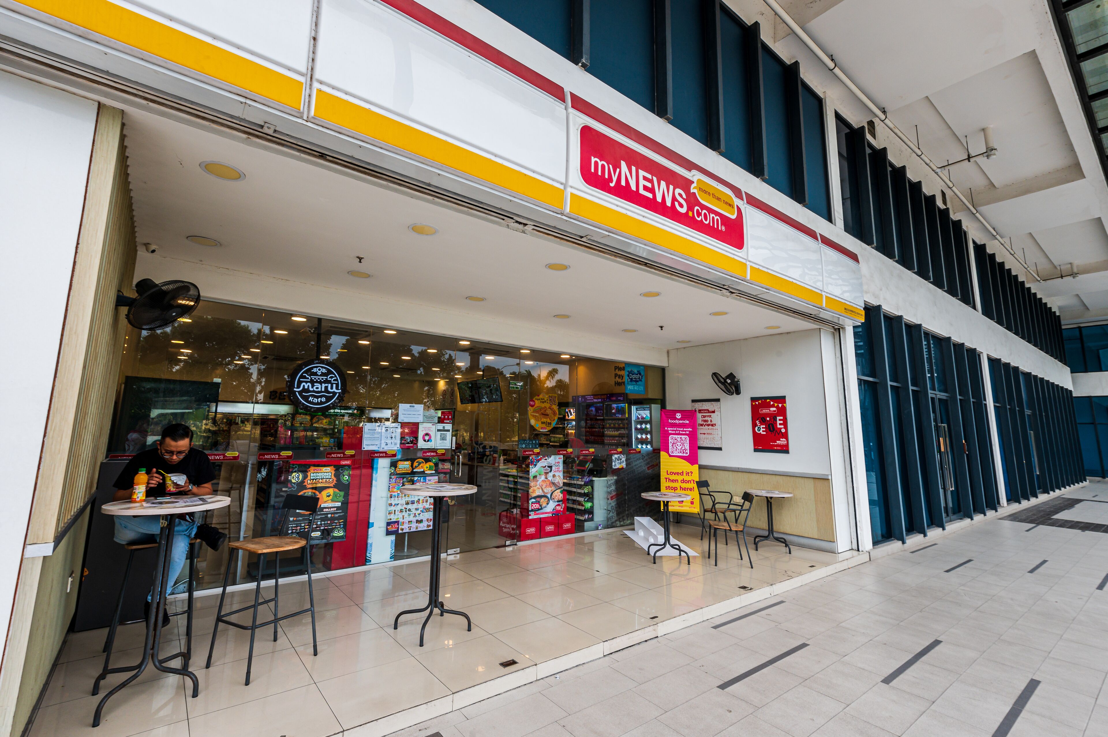 A newsstand with a prominent red and yellow sign, a glass storefront, and a few tables and chairs outside.