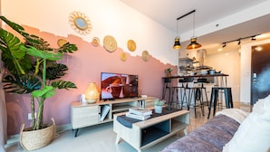 Un salon moderne avec un canapé, une table basse et un téléviseur. On aperçoit un coin repas avec des chaises et, au fond, une cuisine.