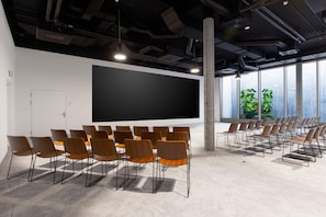A conference room with a large screen, multiple rows of chairs, and large windows with plants. - YOTEL Geneva Lake, Select by Hilton (Founex)