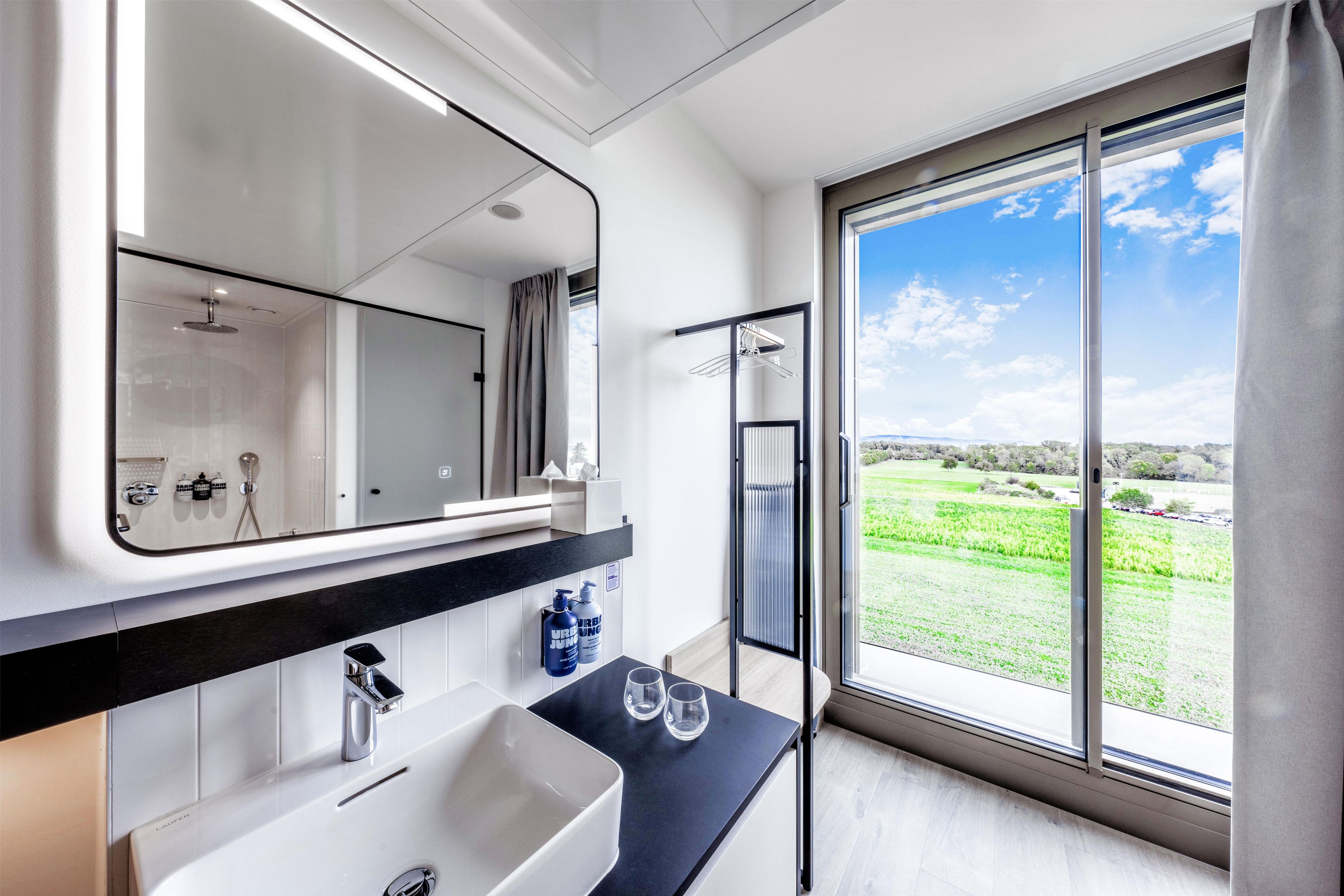 Un baño moderno con un espejo grande, un lavamanos y una ducha. Se observa una ventana con vistas a un paisaje verde.
