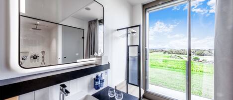 Un baño moderno con un espejo grande, un lavamanos y una ducha. Se observa una ventana con vistas a un paisaje verde.