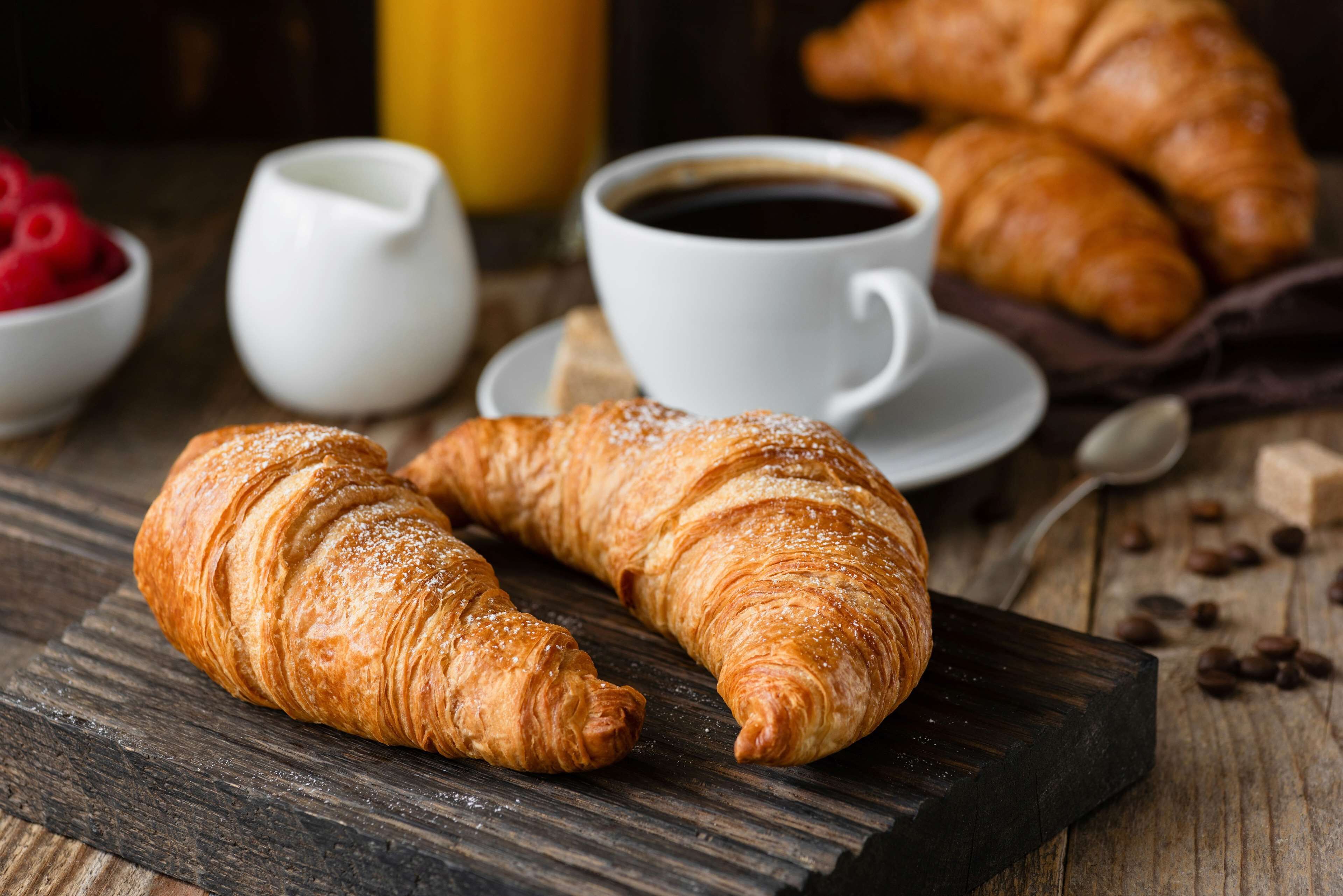 Un croissant sobre una taula de fusta, una tassa de cafè i un bol de mores.