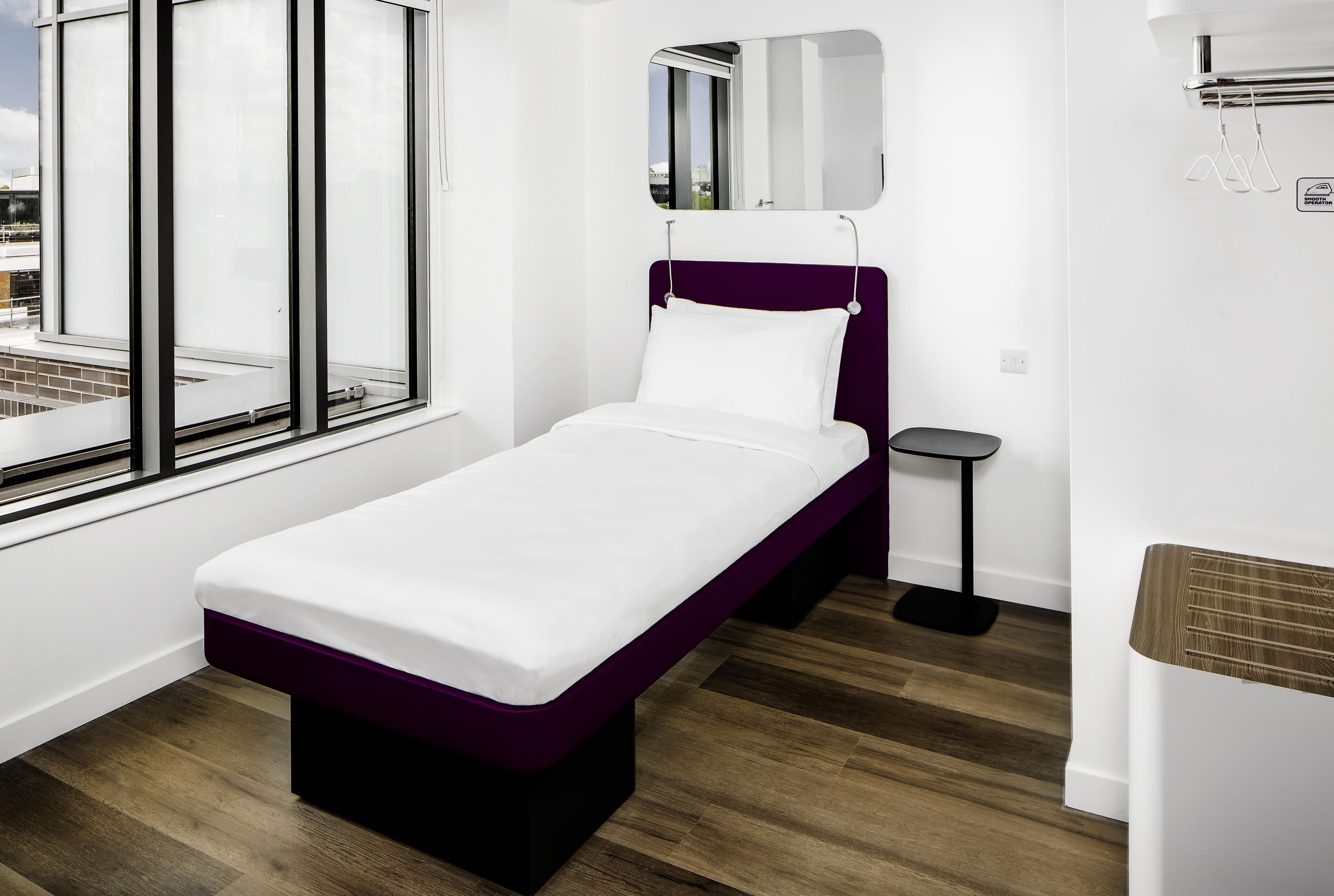 A single-bed room with a window, a bedside table, and a small stool.