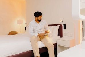 A man sitting on a bed in a hotel room with a white bedspread, a brown headboard, and a bedside table with a lamp.