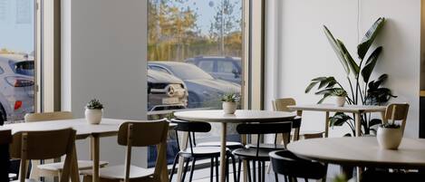 A modern interior with multiple tables and chairs, large windows with a view of parked cars and trees outside.
