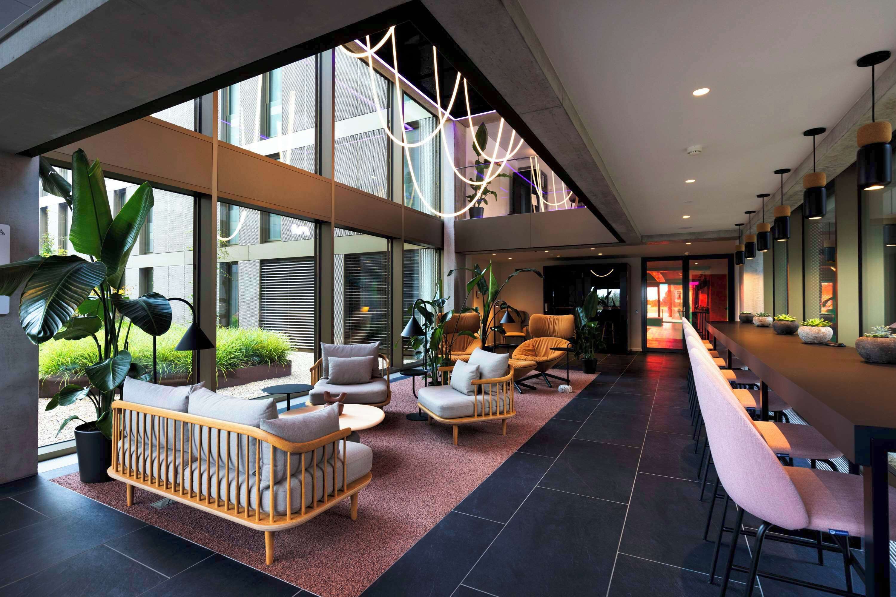 A modern lobby with a long reception desk, seating area with sofas and chairs, large windows, and indoor plants.