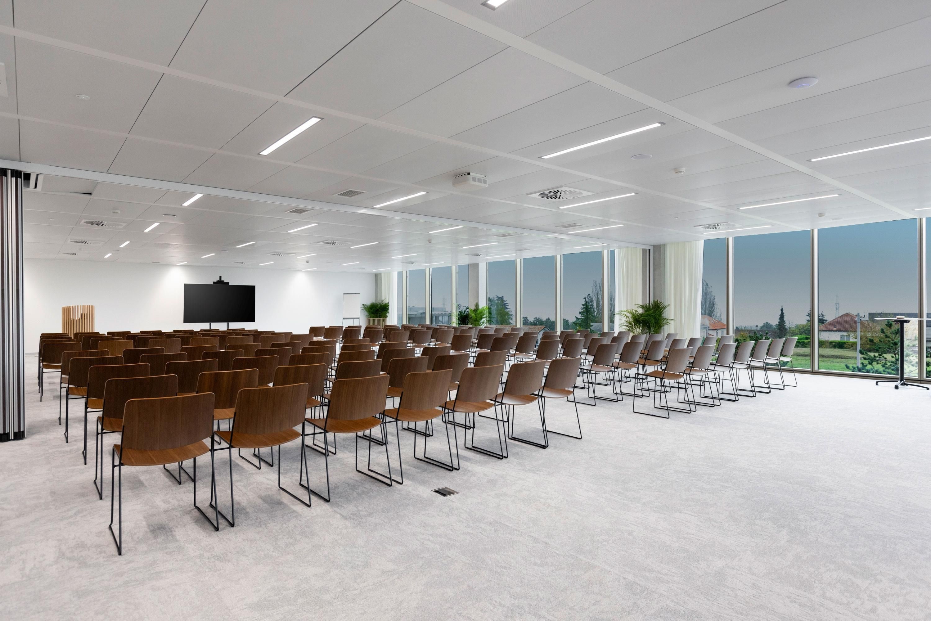 Un salón de conferencias con filas de sillas, una pantalla grande y amplios ventanales que dan a vistas del exterior.