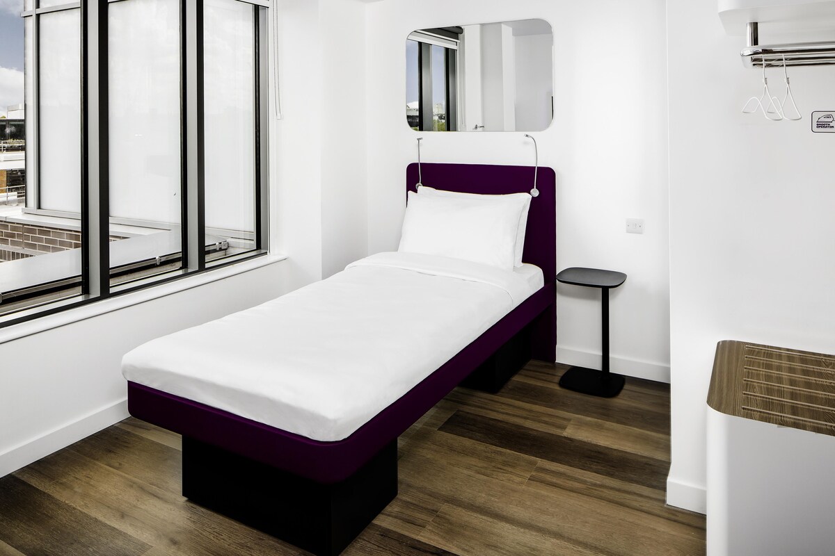 A single-bed room with a window, a bedside table, and a small stool.