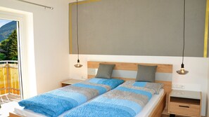 A double bed with a blue and gray patterned bedspread, two bedside tables, a wooden headboard, and a view of a balcony with a wooden fence.