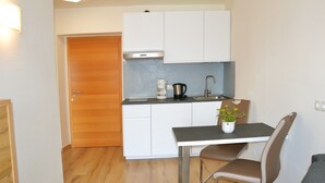 Cocina compacta con cafetera, hervidor y lavamanos. Hay una mesa de comedor con una silla y una planta en maceta.