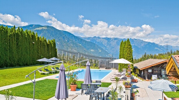 Zona de piscina con sillones de descanso y sombrillas, rodeada de montañas y vegetación.