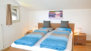Un lit double avec une couvre-lit bleue et grise, deux tables de chevet avec des lampes, une tête de lit en bois, une fenêtre avec des rideaux et un tableau représentant une fleur de lotus accroché au mur.