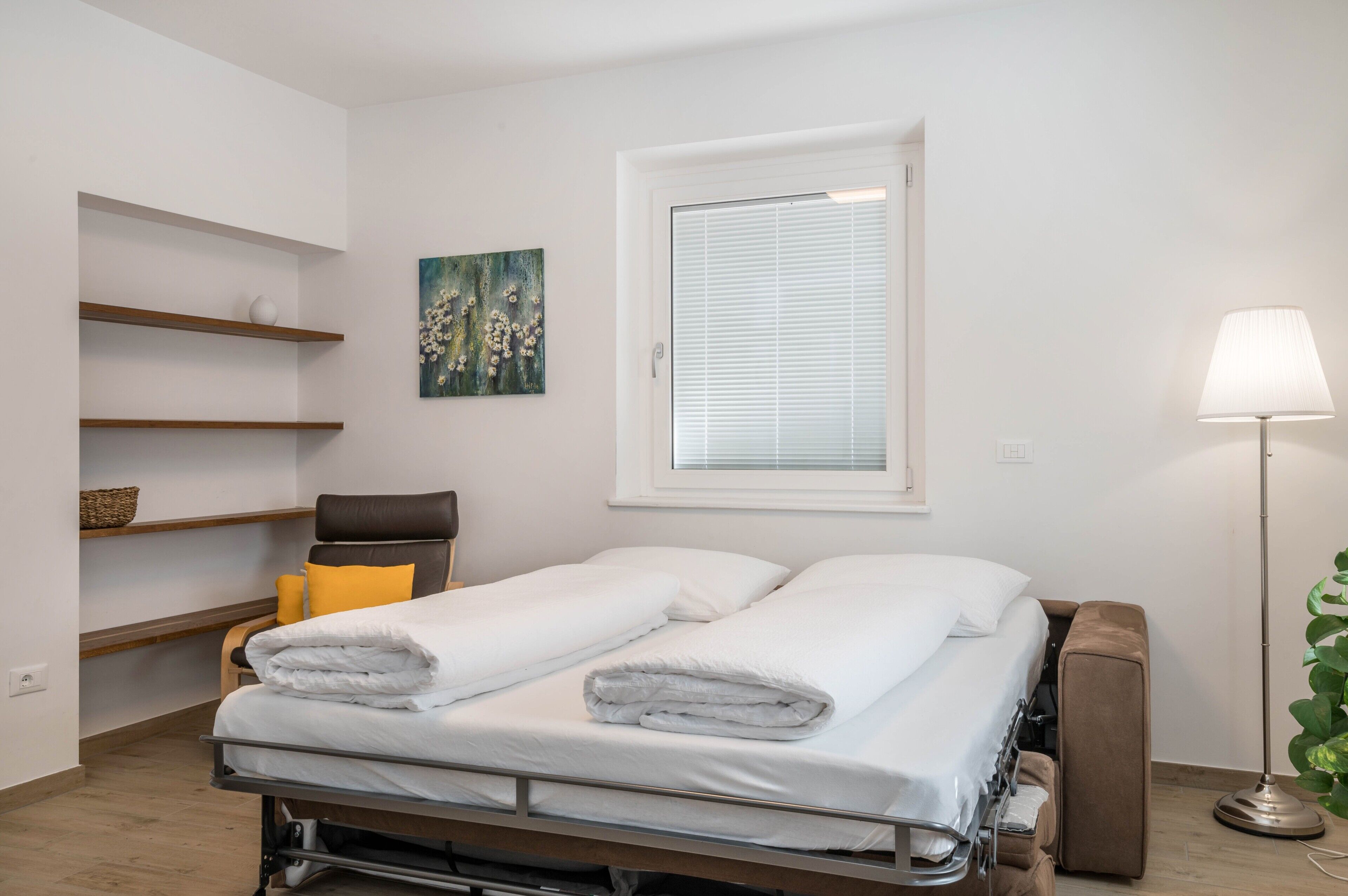 Una cama individual con sábanas blancas, una mesita de noche, una lámpara, una silla, una ventana con persianas y un estante de pared con una cesta.