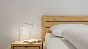 Een houten nachtkastje met een lamp, een open boek en een opgemaakt bed met een wit kussen.