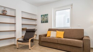 Een woonkamer met een bruine bank, een zwarte stoel, houten planken en een raam met jaloezieën.