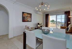 Un coin repas avec une table en verre, des chaises blanches, un grand saladier, un tableau et une vue sur la mer par de grandes fenêtres.
