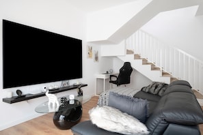 Un salon moderne doté d’un grand téléviseur à écran plat, d’une table basse en verre, d’un canapé en cuir noir, d’un fauteuil de jeu et d’un escalier.