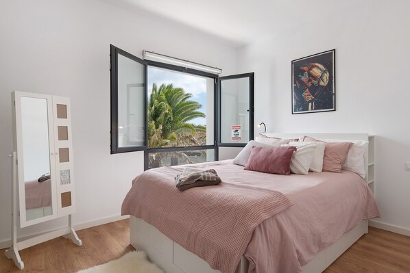 Une chambre avec un lit, une armoire, une fenêtre donnant sur des palmiers et un tableau encadré au mur.