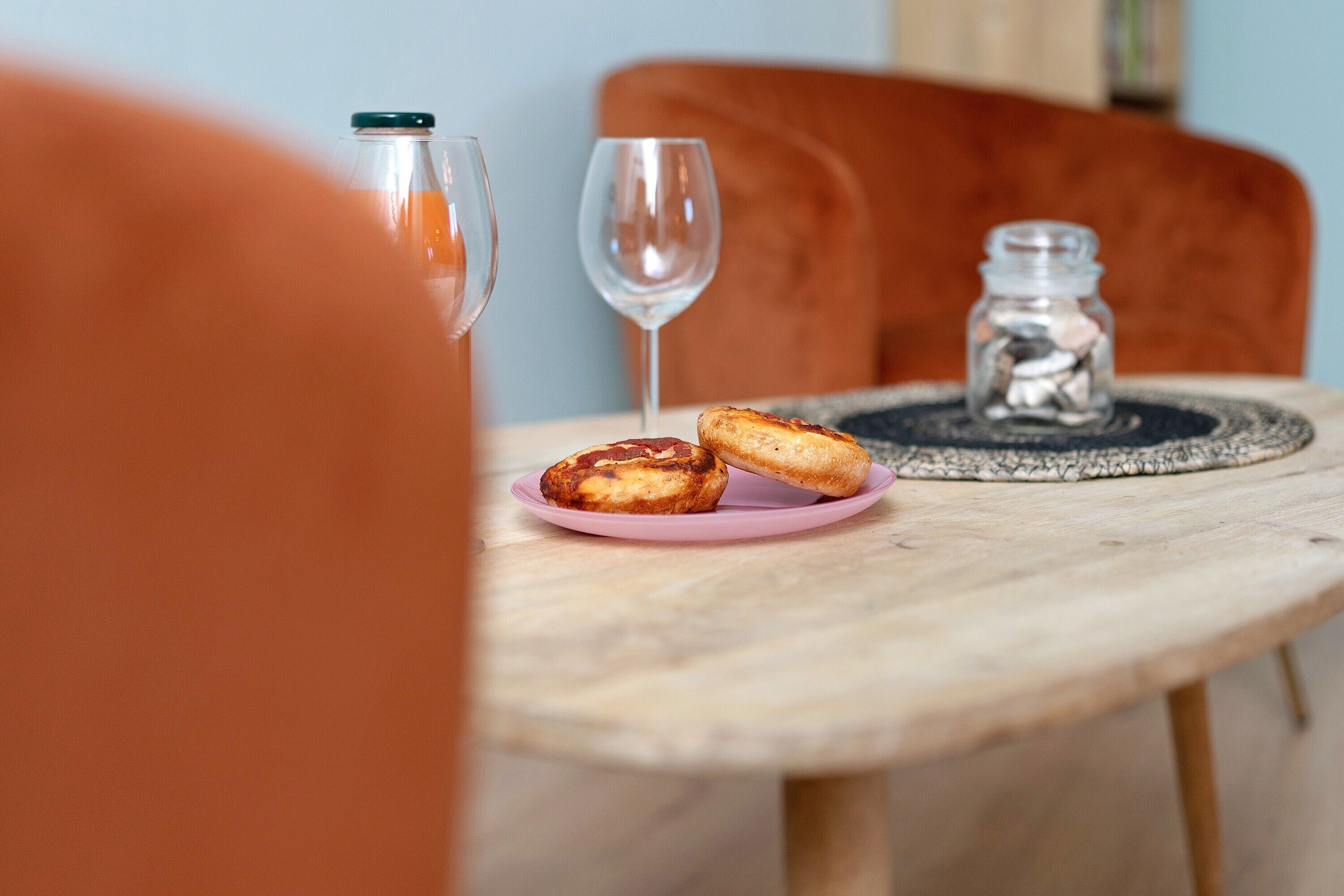 A round wooden table with a pink plate holding two pastries, a clear glass bottle, and a glass wine glass.