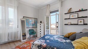 A bedroom with a bed, a desk, a chair, a map on the wall, and a balcony.