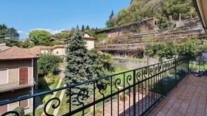 A balcony with a wrought iron railing, a view of a hillside with buildings, and a large evergreen tree.
