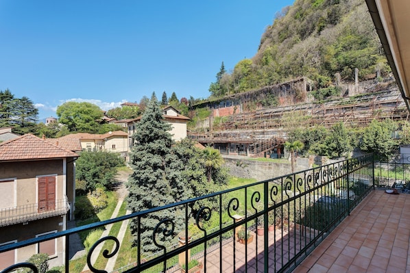 Un balcone con ringhiera in ferro battuto, vista su un pendio con edifici e un grande abete sempreverde.