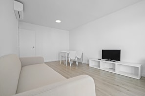 A minimalist living room with a sofa, a TV on a white stand, a small table with chairs, and a door.