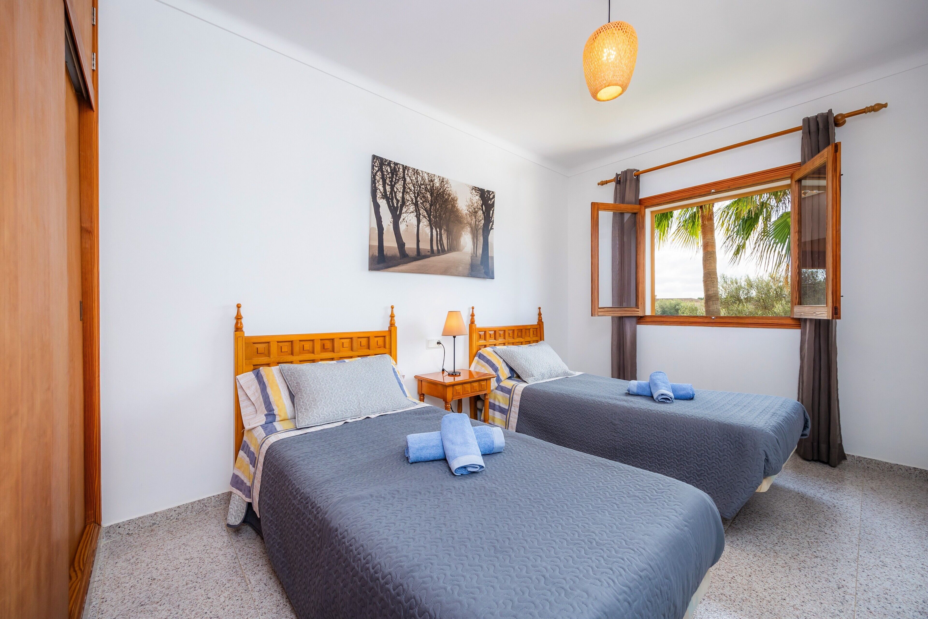 A room with two beds, a wooden headboard, a nightstand, a lamp, a window with curtains, and a painting on the wall.