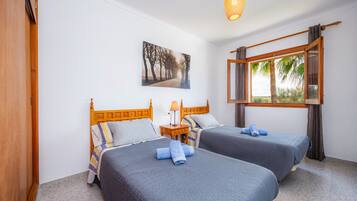 A room with two beds, a wooden headboard, a nightstand, a lamp, a window with curtains, and a painting on the wall.