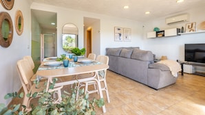 A modern living room with a grey sofa, a dining table with wicker chairs, and a TV. - Holiday Home 'Apt Con Piscina Y Jardín' with Shared Pool, Wi-Fi and Air Conditioning (Puerto Pollensa)