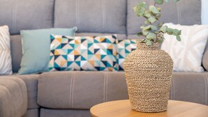 A woven vase with green leaves on a round wooden table in front of a grey sofa with patterned cushions. - Holiday Home 'Apt Con Piscina Y Jardín' with Shared Pool, Wi-Fi and Air Conditioning (Puerto Pollensa)