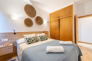 Un letto ben rifatto con copriletto blu e cuscini geometrici, un armadio in legno e decorazioni murali in fibra naturale.