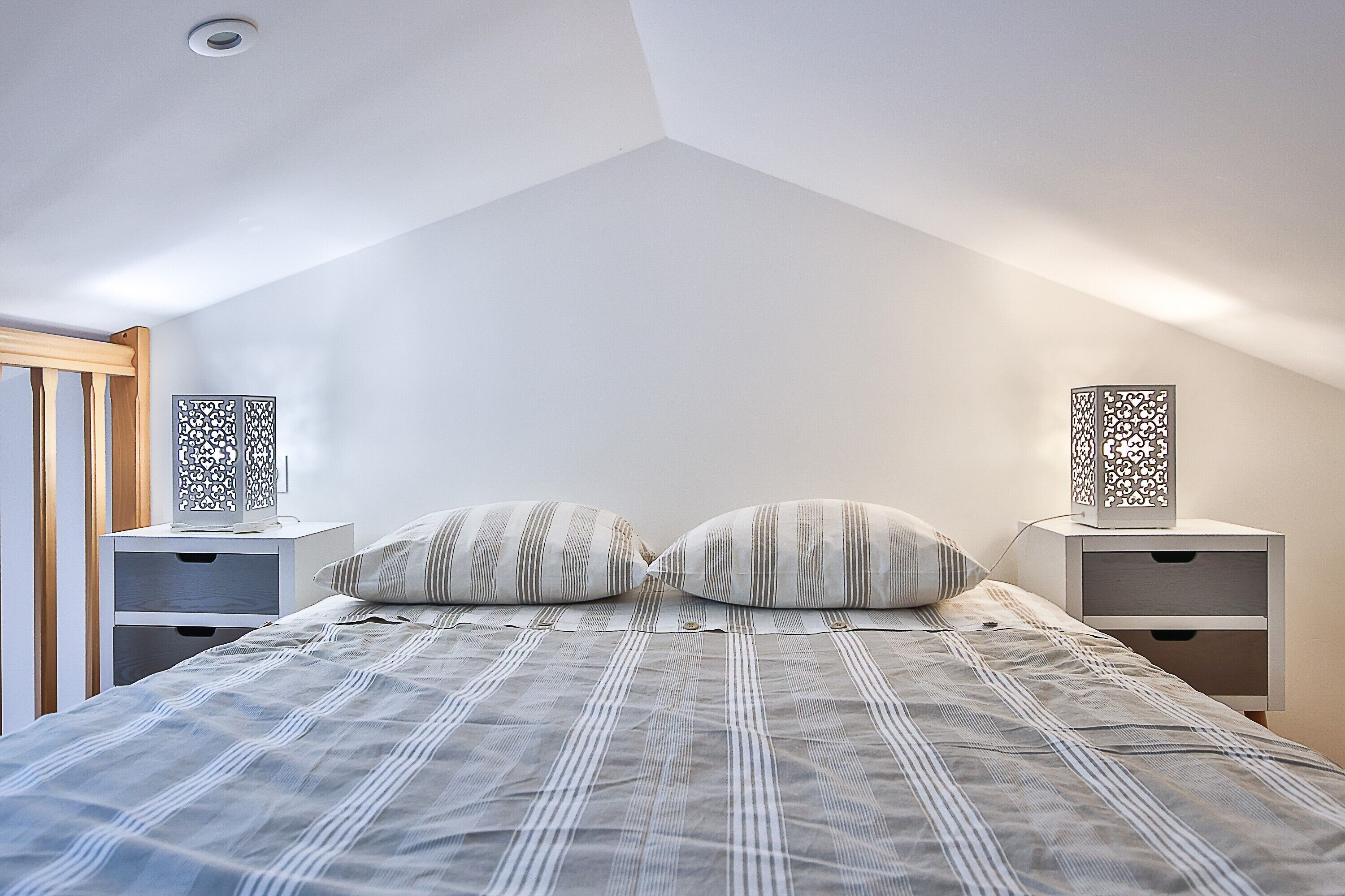 Un dormitorio con techo inclinado, una cama con ropa de cama a rayas, dos mesitas de noche con lámparas decorativas y una puerta de madera.