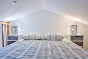 A bedroom with a sloped ceiling, a bed with striped bedding, two bedside tables with decorative lamps, and a wooden door.