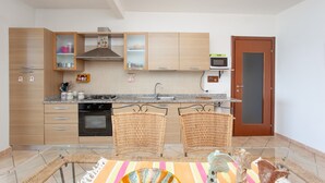A modern kitchen with wooden cabinets, a microwave, an oven, and a dining area with wicker chairs. - Vacation Home Casa sulla Spiaggia 1 with Sea-View, Terraces & Garden (Orosei)