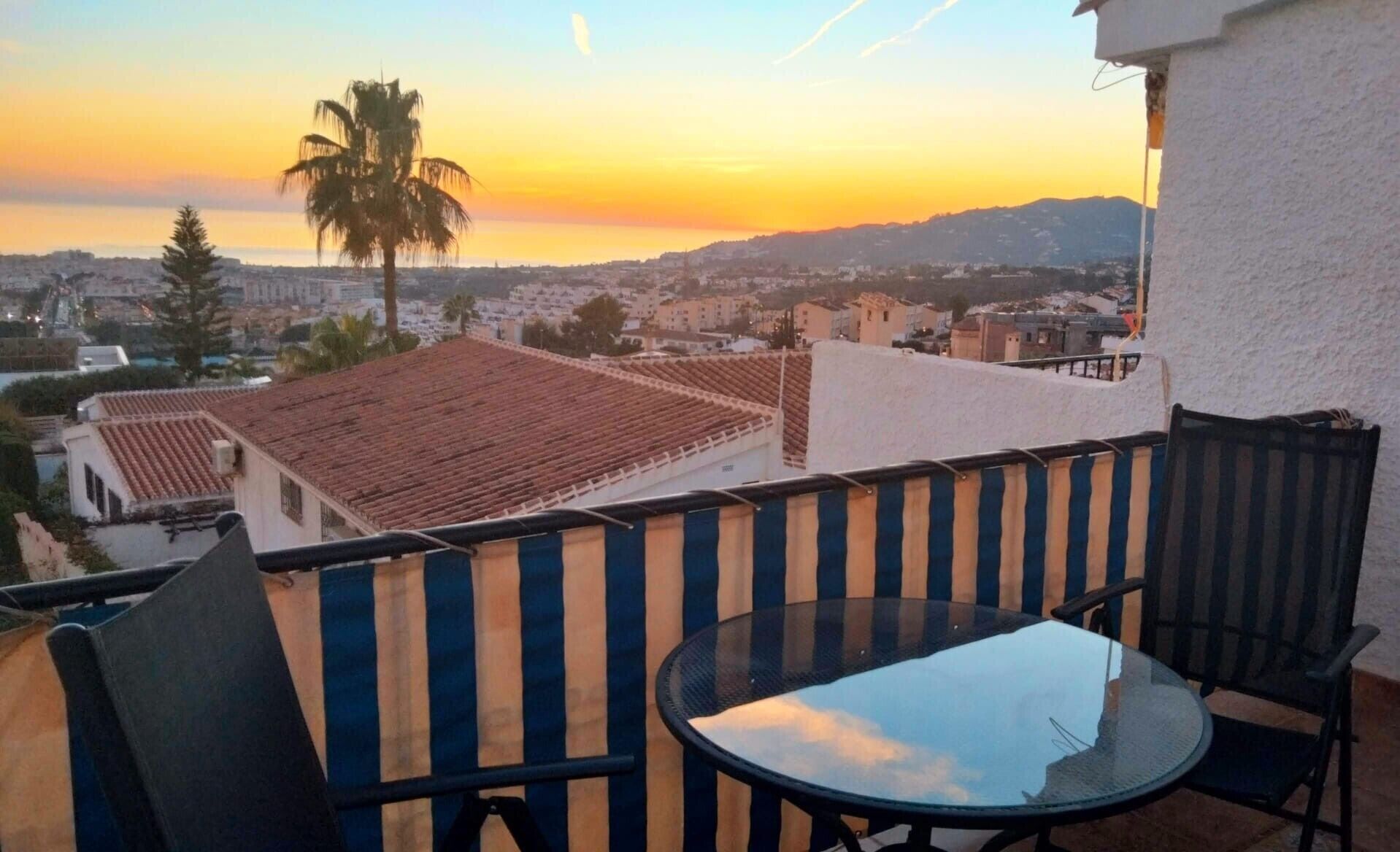 A balcony with a glass table and chairs, overlooking a residential area with terracotta roofs and a palm tree.