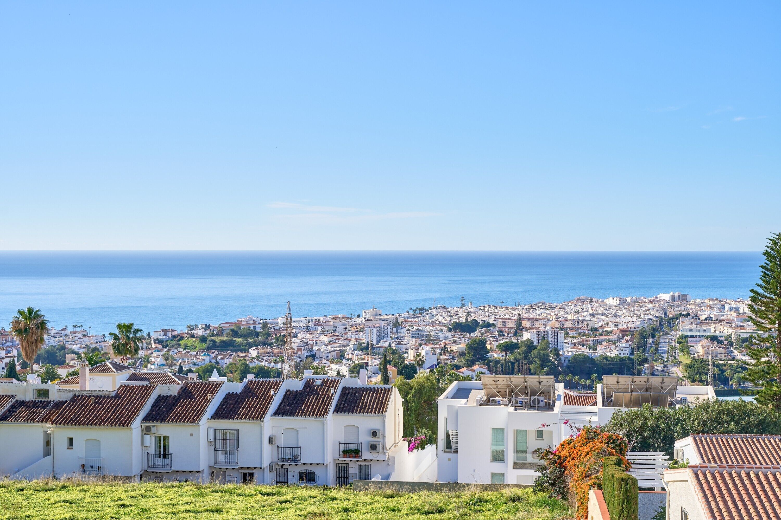 A coastal cityscape with white buildings and terracotta roofs, overlooking the sea.