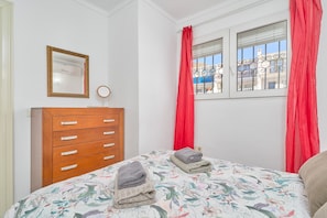 A bedroom with a wooden dresser, a bed with floral bedding, and a window with red curtains. - Apartment 'Los Robles ' with Sea View, Wi-Fi and Air Conditioning (Nerja)