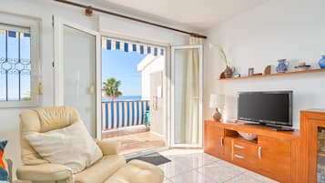 Un salón con sofá, televisión, mueble de madera y vistas al mar a través de un ventanal abierto.