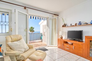 A living room with a sofa, TV, wooden cabinet, and a view of the sea through an open door. - Apartment 'Los Robles ' with Sea View, Wi-Fi and Air Conditioning (Nerja)
