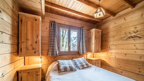 Quarto de cabana de madeira com uma cama, duas mesas de cabeceira, uma janela com cortinas e um lustre.