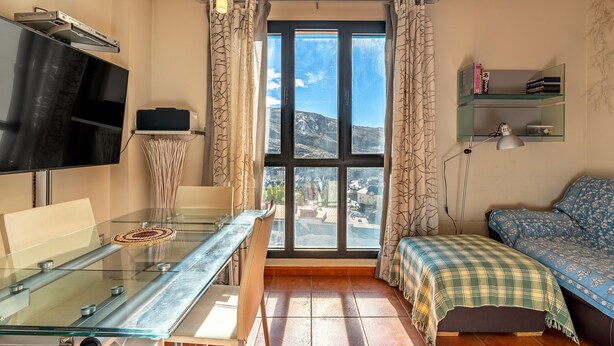 A compact living space with a glass dining table, a sofa, a TV, and a large window with curtains. - Apartment 'Diseño A Pie De Pista' with Mountain View and Wi-Fi (Monachil)